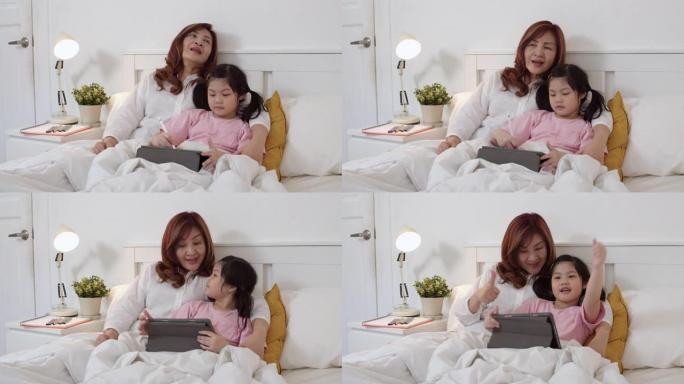 亚洲祖母用平板电脑在家给孙女读童话故事。高年级中国人，奶奶开心放松年轻女孩睡前躺在床上晚上在家卧室的