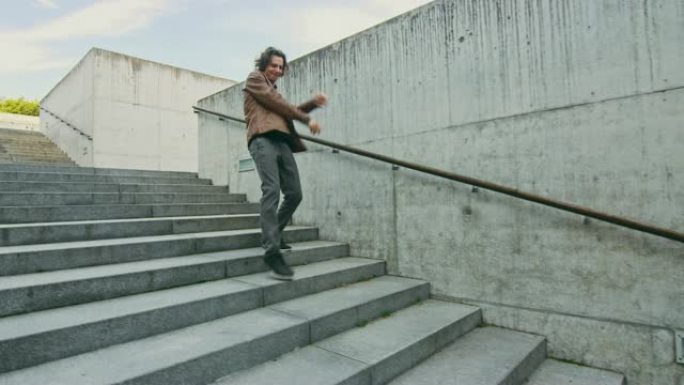 开朗快乐的长发年轻人在走下楼梯时积极跳舞。他穿着一件棕色皮夹克。在城市混凝土公园拍摄的场景。天很亮。