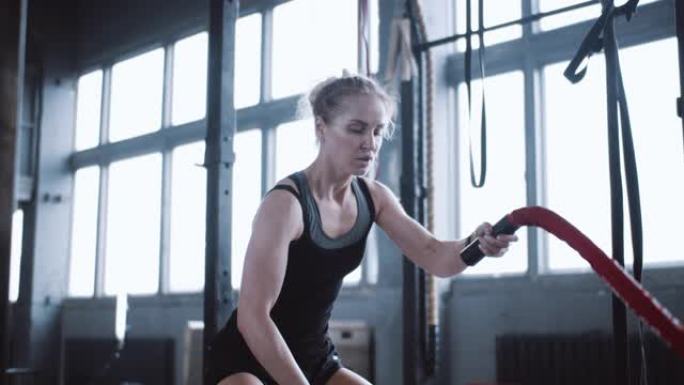 年轻美丽坚定的运动金发女子用战斗绳锻炼，在大型健身房进行功能性训练锻炼。