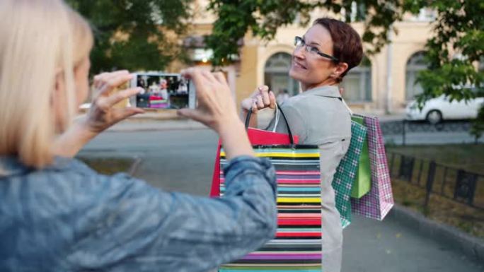 迷人的女人带着购物袋在户外为智能手机相机摆姿势