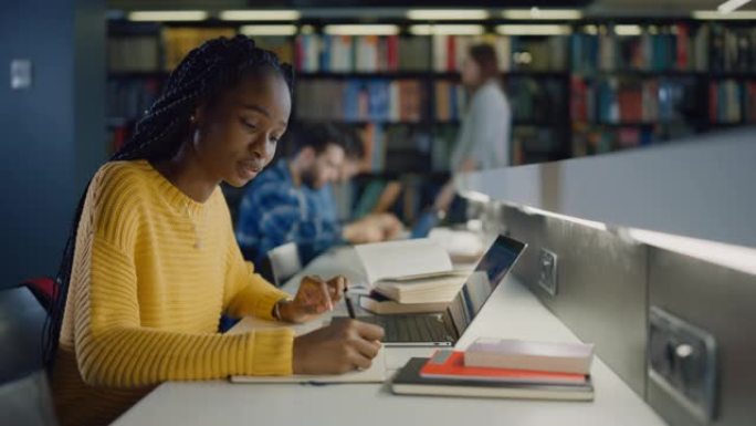 大学图书馆: 有天赋的黑人女孩使用笔记本电脑，为论文，论文写笔记，为课堂作业学习。多样化的多民族学生