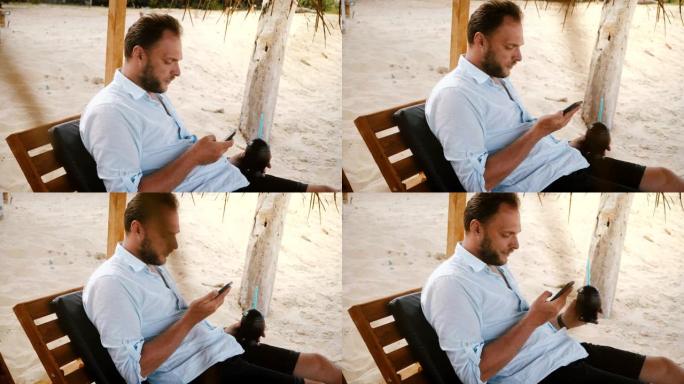 快乐自信的男性游客，智能手表和水果鸡尾酒坐在沙滩椅上，使用智能手机社交网络