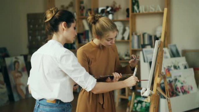 勤奋的学生在现代工作室的专业艺术家的指导下绘画。女人指着画布并提供建议。