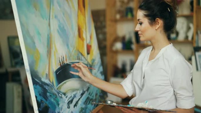 微笑的年轻女子正在用油画在画布上描绘船和蓝色海浪的画面，她拿着画笔和调色板站在画架旁。