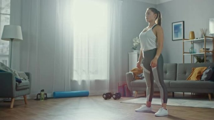 穿着运动服的强壮美丽的运动健身女性正在她明亮宽敞的客厅做蹲下锻炼，室内简约。