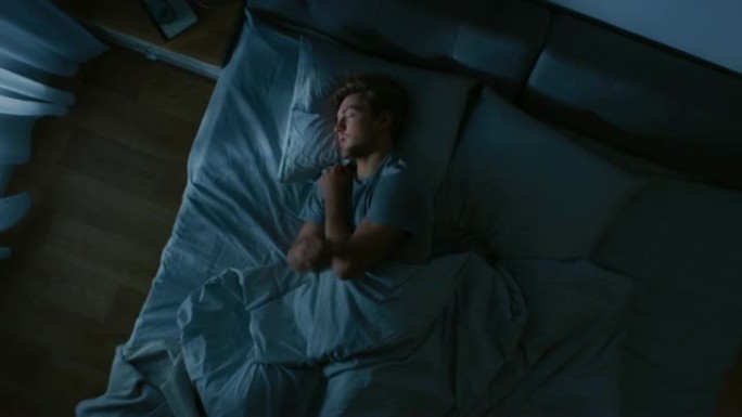 英俊的年轻人晚上舒适地睡在卧室的床上的俯视图。蓝色夜间颜色，寒冷微弱的灯柱光线透过窗户照射。