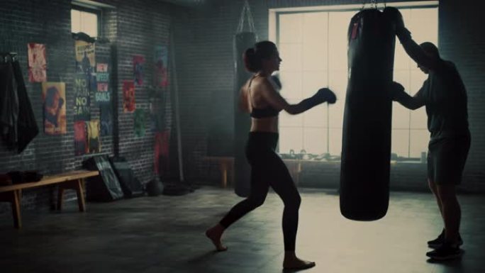 健身运动女性跆拳道运动员在阁楼健身房锻炼时拳打出气筒。她美丽而充满活力。强壮的教练拿着拳击包。激烈的