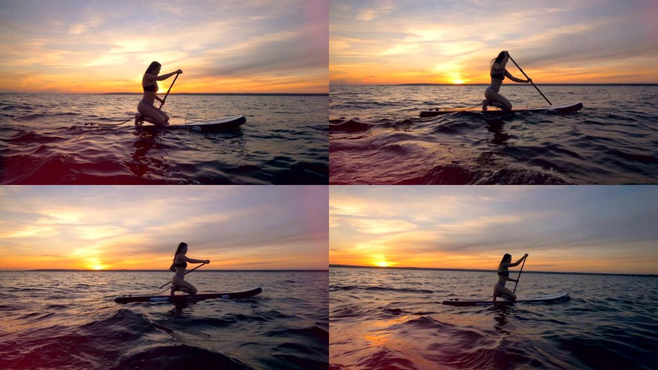 一个女人在日落背景上划船，侧视图。一个女孩骑着她的冲浪板，在海里。
