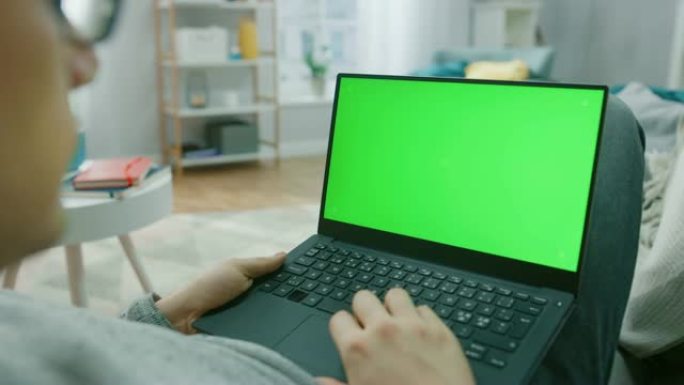 在家中的男人坐在沙发上，在绿色模拟屏幕笔记本电脑上工作。家伙使用笔记本电脑设备，浏览互联网，观看内容