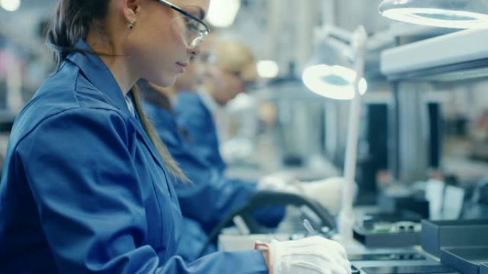穿着蓝色工作服和防护眼镜的女电子工厂工人正在为带有镊子的智能手机组装印刷电路板。背景员工更多的高科技