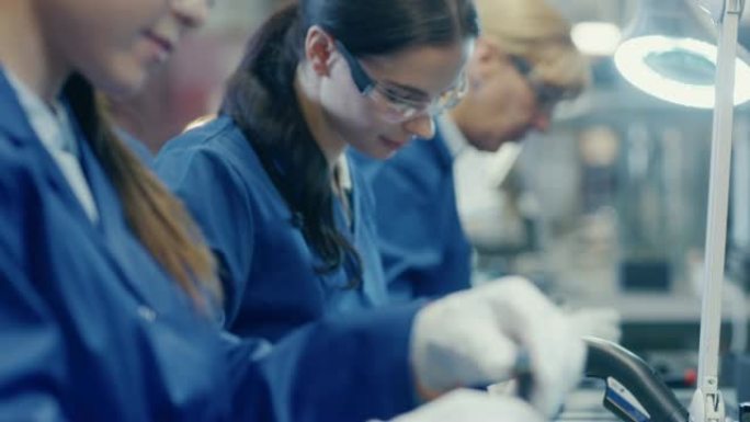 穿着蓝色工作服和防护眼镜的电子工厂女工人正在用螺丝刀组装智能手机。高科技工厂设施，后台有更多员工。