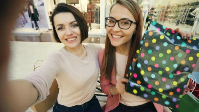 漂亮的女孩朋友在购物中心用纸袋自拍，开心地展示舌头和做有趣的脸。友谊和技术概念。