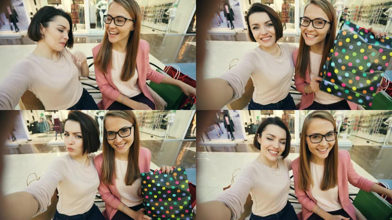 漂亮的女孩朋友在购物中心用纸袋自拍，开心地展示舌头和做有趣的脸。友谊和技术概念。