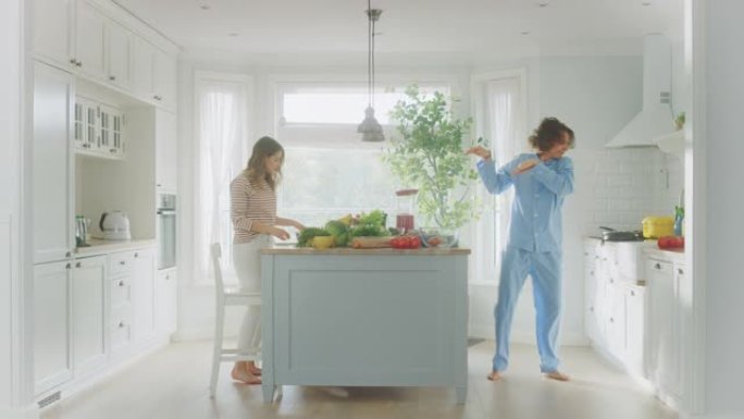 穿着蓝色睡衣的英俊年轻人和漂亮的女孩在厨房里跳舞。女性使用智能手机，从长发的男性那里捕获绿色蔬菜。晴