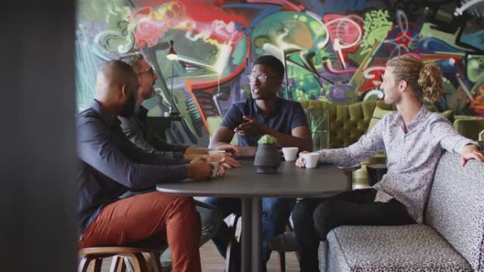 四位男性创意同事在工作场所食堂的一张桌子上聊天，四分之三
