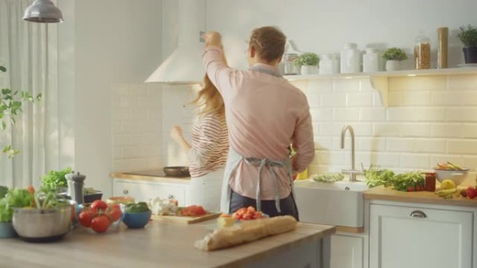 穿着粉色衬衫和围裙的英俊年轻人和穿着条纹套头衫的漂亮女孩在厨房里创造性地跳舞。阳光明媚的现代厨房，桌