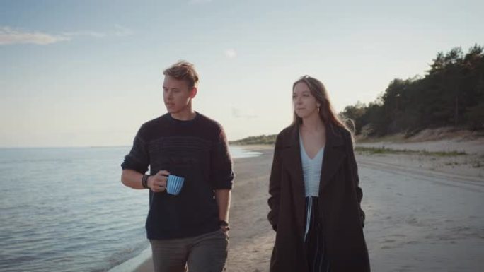 年轻夫妇在海滩上散步。多云温暖的日子里平静的海面。帅哥拿着茶杯。漂亮的女孩穿着外套。大自然中浪漫的夏