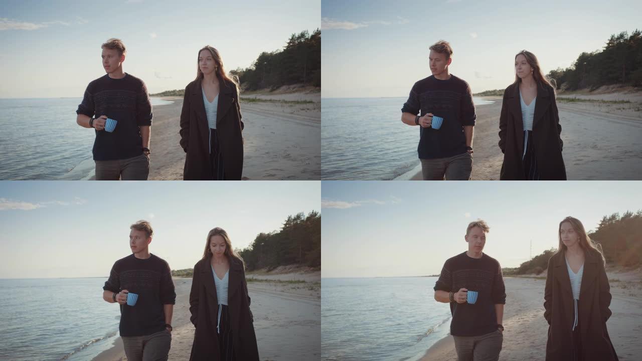 年轻夫妇在海滩上散步。多云温暖的日子里平静的海面。帅哥拿着茶杯。漂亮的女孩穿着外套。大自然中浪漫的夏