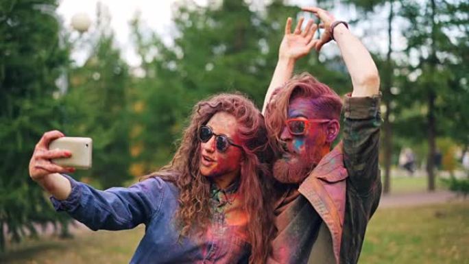 脸上和头发被油漆覆盖的女孩和男孩正在与智能手机一起自拍，站在公园的户外，享受派对和技术的乐趣。