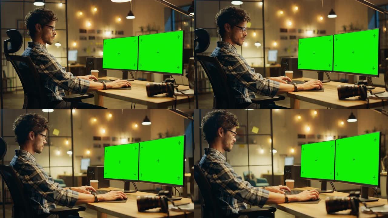 傍晚在创意办公室: 专业摄影师在带有两个绿色模拟屏幕的台式计算机上工作。带悬挂灯泡的现代工作室办公室