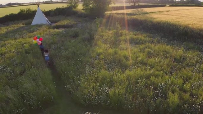 在音乐节上露营的两个女性朋友的空中无人机拍摄，穿过田野朝着拿着气球的圆锥形帐篷奔跑-慢动作射击