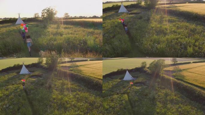 在音乐节上露营的两个女性朋友的空中无人机拍摄，穿过田野朝着拿着气球的圆锥形帐篷奔跑-慢动作射击