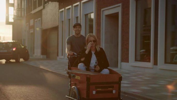 英俊的年轻人骑着成人三轮车，女友坐在前装载拖车上。热恋中的年轻夫妇一起骑自行车穿过时尚的城市街道