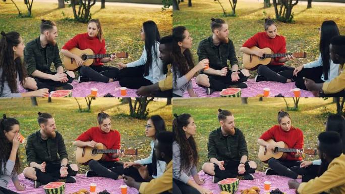 多种族的朋友正在弹吉他，唱歌和享受饮料，坐在公园草坪上的格子上，享受休闲和陪伴。