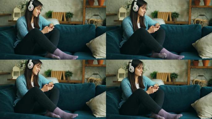 漂亮的女生正在听音乐，戴着耳机，触摸智能手机屏幕，坐在沙发上在家玩得开心。娱乐和休息概念。