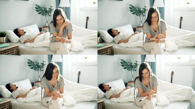 紧张的女孩坐在床上拿着怀孕测试，而男孩在卧室睡觉