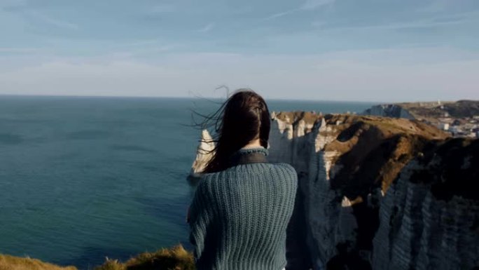 背景拍摄，兴奋的女游客看着华丽的埃特雷塔岩石和海洋全景，头发在风中吹动。