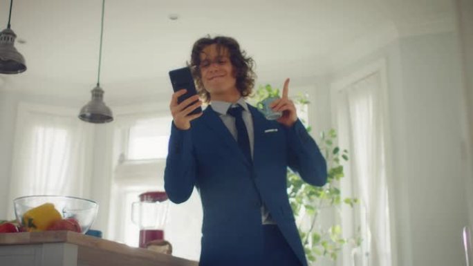 穿着蓝色西装的长发快乐的年轻人在家里跳舞。他正在手机上听音乐。精力充沛的人在厨房里使用智能手机，里面
