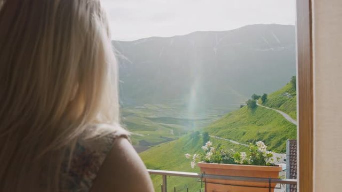 年轻女子女士分开酒店窗帘，享受阳光明媚的田园诗般的山景，意大利翁布里亚城堡