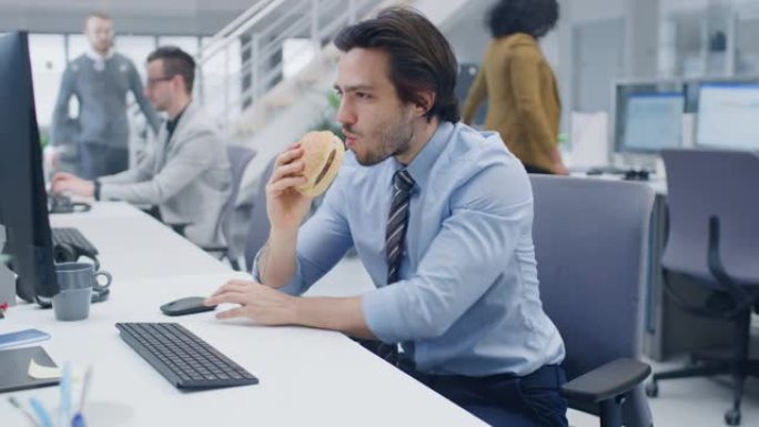 在明亮的现代办公室里，年轻的商人坐在台式计算机上吃汉堡包，他生气地咬着面包，并在午餐期间继续工作。在