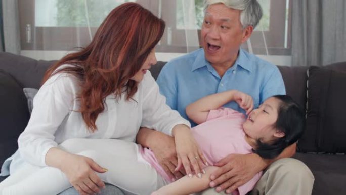 亚洲祖父母在家和孙女聊天。中国老年人，老一代，祖父和祖母的家庭时间与年轻的女孩孩子在客厅的沙发上放松