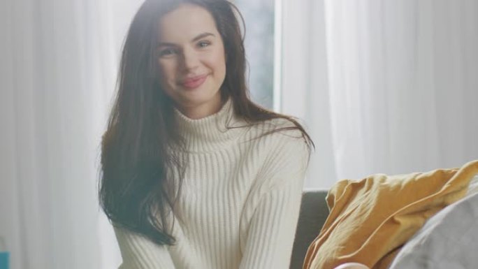 美丽的年轻女子坐在椅子上对着镜头迷人地微笑。周日早上，穿着超大号白色针织毛衣的害羞性感女孩在舒适的公