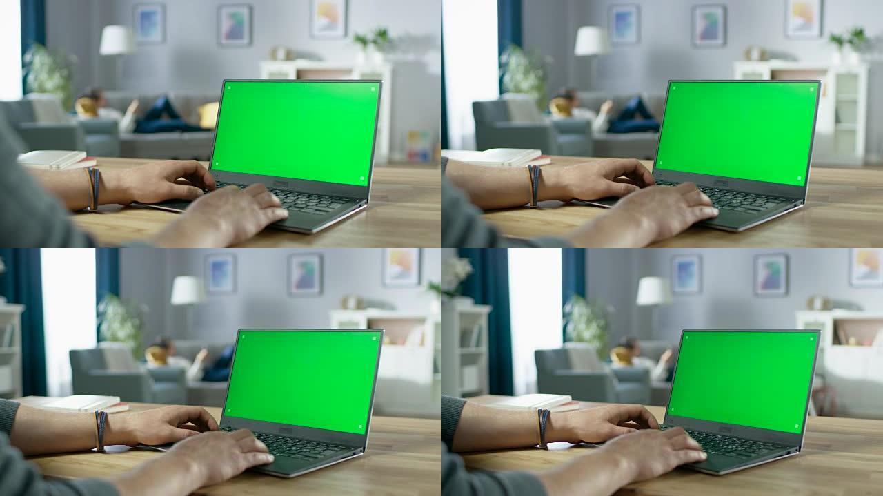 在带有绿色模拟屏幕的笔记本电脑上，男人的手在肩膀上打字。在背景舒适的客厅，注意力不集中的女人躺在沙发