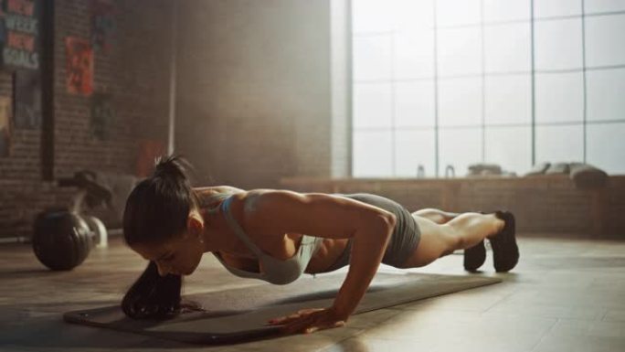 穿着运动上衣和短裤的强壮而健康的运动女性正在阁楼风格的工业健身房进行俯卧撑运动，并配有励志海报。这是
