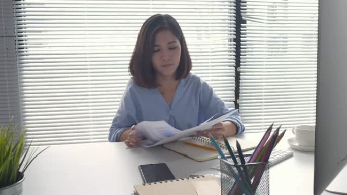 美丽的年轻应变亚洲妇女坐在家里的客厅里在笔记本电脑上工作。在家庭办公室工作的亚洲女商人。