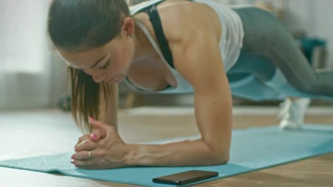 穿着运动运动服的强壮美丽的健身女孩正在用手机上的秒表做木板运动。她正在室内舒适的客厅里训练。