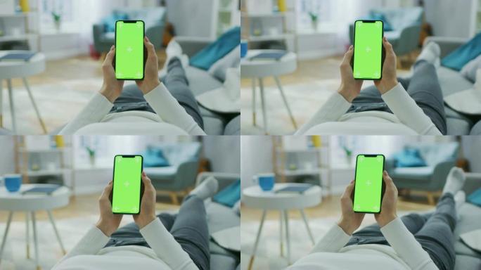 家里的年轻女子躺在沙发上，使用绿色模拟屏幕智能手机。女孩使用色键手机，浏览互联网，观看内容。视点摄像