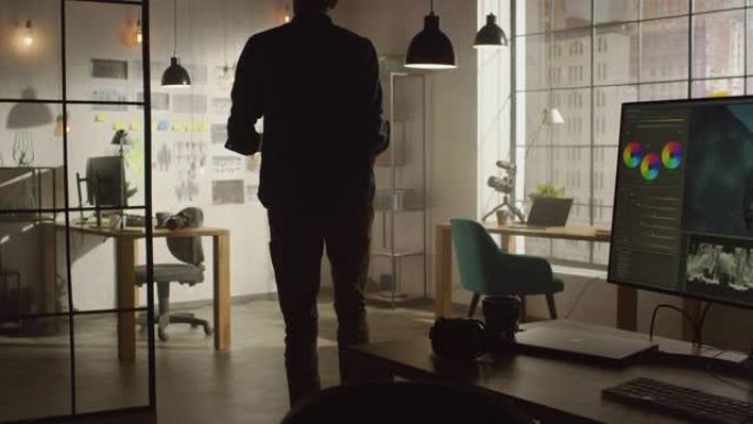 年轻的有创造力的人站在他的桌子上，用个人电脑和视频编辑软件显示，并在他凉爽的阁楼办公室里用有组织的故