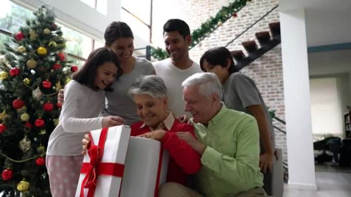 兴奋的祖母打开一个大圣诞礼物，而充满爱心的家人在她的微笑中