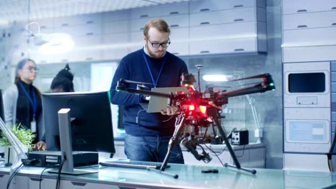 年轻的男性工程师在手拿笔记本电脑时对无人机进行了编程。他在一个明亮的现代高科技实验室工作。