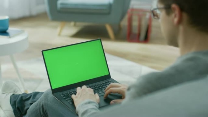 家里的年轻人在带有绿色模拟屏幕的笔记本电脑上工作。他坐在舒适客厅的沙发上。肩膀上的镜头。