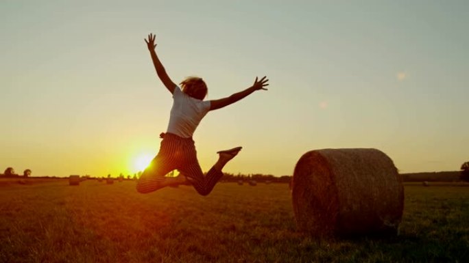 慢动作。女士无忧无虑的女人在日落时带着干草在田园诗般的乡村田野中跳跃