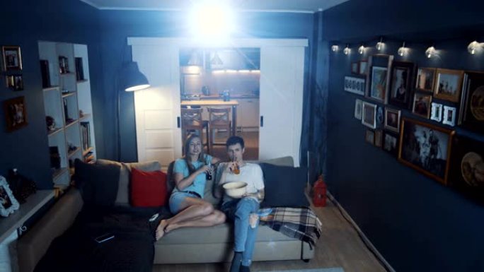快乐的年轻高加索夫妇在家使用投影仪和大屏幕在电视上看电影，网络在线电影慢动作。