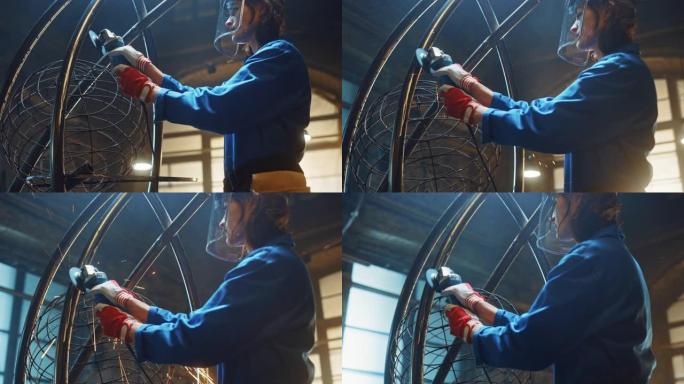 穿着蓝色连身裤的年轻当代女艺术家正在工作室里用角磨机打磨一个金属管雕塑。赋权女人用金属制作现代残酷的