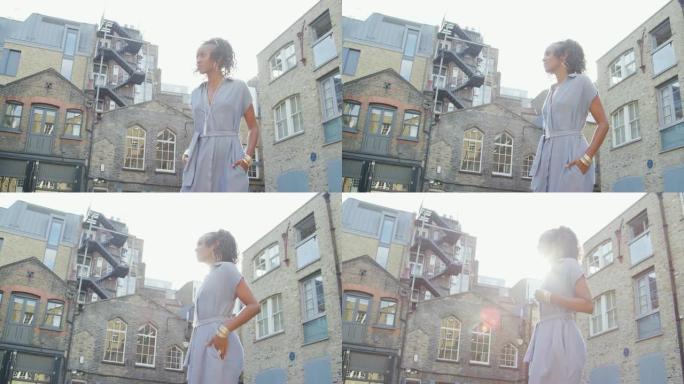 穿着蓝色连衣裙的时尚年轻黑人女性站在阳光下对着建筑物，镜头耀斑，低角度