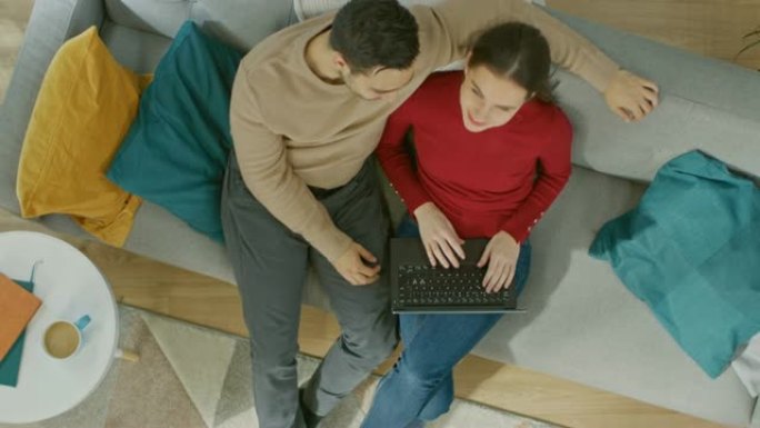 年轻美丽的夫妇坐在沙发上，用笔记本电脑。他们看着上面微笑。舒适的客厅，配有现代室内植物、桌子和木地板